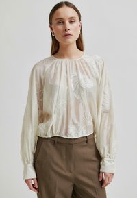 Könnyű, krémszínű blúz hosszú ujjakkal, ráncolt szegéllyel és virágmintás hímzéssel. Elegáns barna nadrággal párosítva.