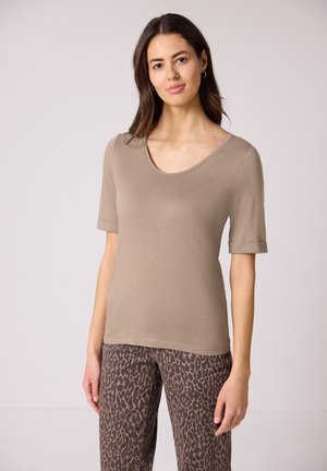 Woman wearing taupe scoop-neck short sleeve top and brown leopard print pants, standing against a plain light background.