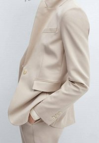 Béžový na míru šitý blazer, zapínání na jeden knoflík, klopy s výřezem, dva přední kapsy, zdůrazněné švy, hladká textura a manžety na knoflíky.