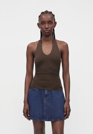 Femme aux cheveux tressés portant un haut marron à col halter et une jupe en denim bleu, debout devant un fond blanc uni.