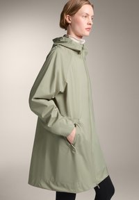 Jachetă waterproof de culoare verde deschis, cu glugă, dotată cu buzunare laterale și un croi lejer. Textură netedă și design ușor pentru confort.