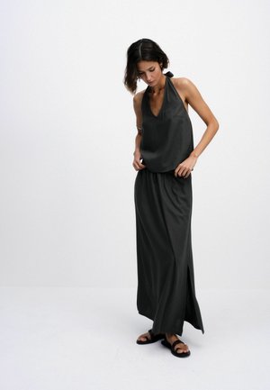 Woman adjusting sleeveless black halter top and matching long skirt with side slit, wearing black sandals, standing against white background.