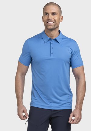 Lächelnder Mann, der ein blaues kurzärmeliges Poloshirt und dunkle Hose trägt, steht mit entspannt an den Seiten hängenden Händen vor einem schlichten Hintergrund.