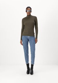 Olive ribbed turtleneck sweater paired with light blue jeans and black ankle boots. Model wears mirrored sunglasses, standing against a white background.