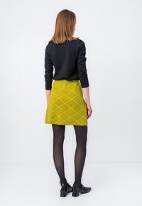Kollane tepitud mini-seelik, millel on teemantmustrit, kombineerituna musta pikakäistega pluusi ja mustade retuusidega, nähtuna tagant.