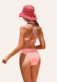 Bikini de cuadros rosa con tirantes entrecruzados y detalle de lazo en la espalda, combinado con un sombrero de cubo de rayas rojas y blancas. Textura suave en todo.