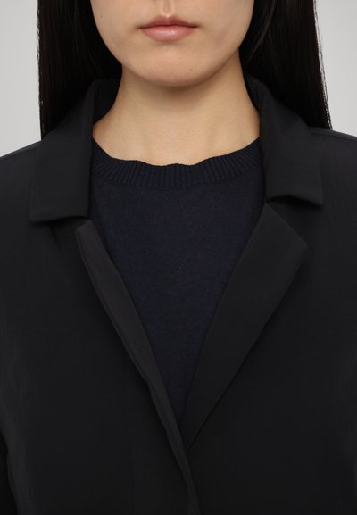 Čierny, ľahký blazer s klopami nad tmavomodrým rebrovaným sveterom. Hladká textúra s minimálnymi detailmi.