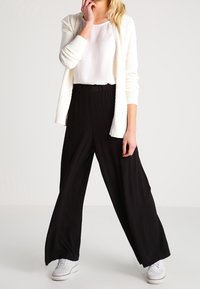 Vit stickad cardigan över en vit topp, i kombination med svarta vida byxor och vita sneakers. Outfiten har en avslappnad passform och mjuka texturer.