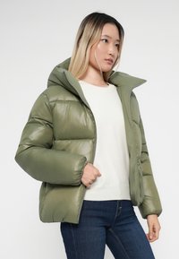 Giacca imbottita verde oliva con colletto alto, chiusura a zip e polsini elasticizzati. Presenta una finitura lucida e una forma squadrata. Indossata sopra una maglietta leggera.