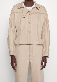 Veste en denim beige avec un col, des boutons sur le devant et des accents de couture jaune. Présente un design court et une coupe décontractée.