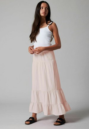 Ung kvinne med langt mørkt hår, iført hvit singlet, lys rosa lagdelt maxiskjørt og svarte sandaler, stående mot en grå bakgrunn.