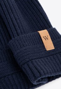 Gorro de punto azul marino hecho de tela suave y texturizada con costillas verticales. Presenta una etiqueta rectangular de cuero marrón con logo en relieve.