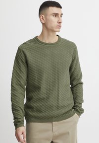 Groene gebreide trui met een gestructureerd, rasterachtig patroon, ribbelkraag en lange mouwen, gecombineerd met beige broeken.