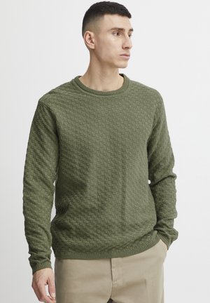 Groene gebreide trui met een gestructureerd, rasterachtig patroon, ribbelkraag en lange mouwen, gecombineerd met beige broeken.