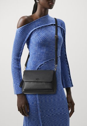 Femme portant une robe bleu côtelée à une épaule avec des manches longues et portant un sac bandoulière en cuir noir avec une fermeture à rabat.