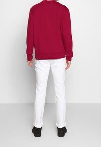 Rode sweatshirt met lange mouwen, geribbelde manchetten en zoom, gecombineerd met witte jeans en zwarte schoenen, gezien van achteren. Glad oppervlak.