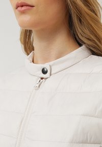 Närbild av en kvinnas nacke och haka som bär en ljusbeige vadderad jacka med dragkedja och knappad ståkrage.