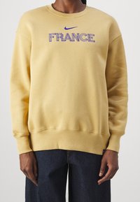 Osoba ubrana w jasnożółtą bluzę z napisem "FRANCJA" i niebieskim logo Nike wyszytym na piersi, w połączeniu z ciemnoniebieskimi dżinsami.