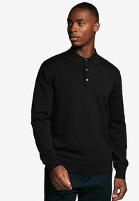 Melna ilga piedurkne polo krekls ar apkakles izgriezumu, trīs pogu aizdari un ribotas aproces, uz vienkārša fona.