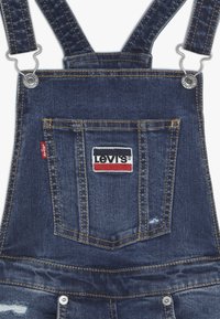 Salopette in denim con spalline blu regolabili, hardware argentato e una tasca frontale con etichetta rossa e bianca di Levi's. Dettagli strappati presenti.