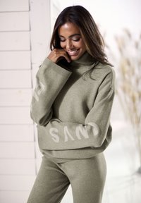 Hellgrüner Strickpullover mit Rollkragen und überschnittenen Schultern, ausgestattet mit weißen "SAND"-Text auf den Ärmeln, kombiniert mit passenden schmalen Hosen.