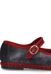 Zapato de lona tipo slip-on con ribete rojo y correa con un acento dorado circular. Textura entrelazada alrededor de la base en un color contrastante.
