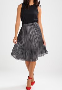 Czarny top bez rękawów w połączeniu z plisowaną, metalicznie srebrną spódnicą midi oraz czerwonymi szpilkami, podkreślający dopasowaną talię i płynny materiał.