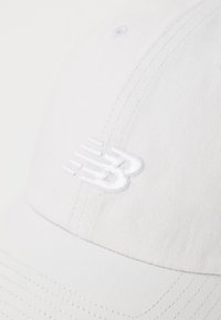 Căciulă de baseball albă, realizată dintr-un material texturat, cu un logo alb cusut în față. Vârful este curbat, cu detalii vizibile ale cusăturii.