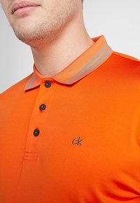Polo naranja con un cierre de dos botones, botones negros y un cuello a rayas grises. Presenta un pequeño logotipo bordado "ck" en el pecho.