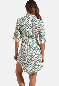 Korte mouwen jurk in roomkleur met een blauw geometrisch bladpatroon. Heeft een ceinture taille en zijsplitten voor bewegingsgemak.