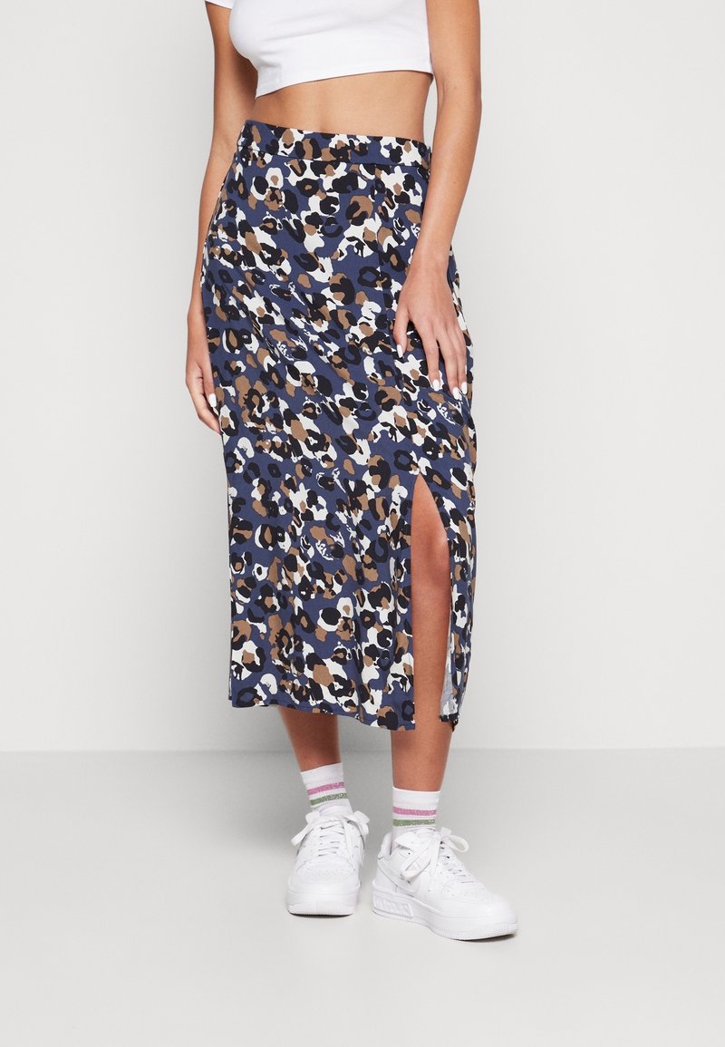 Femme portant une jupe midi à imprimé léopard bleu, marron, noir et blanc avec une fente devant, un crop top blanc, des baskets blanches et des chaussettes rayées.