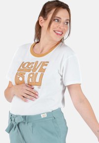 Weißes T-Shirt mit einem runden Senfgelb-Kragen und braunem "LOVE IS ALL" Grafiktext. Kombiniert mit tealten, hochgeschnittenen Hosen mit einer Schleifendetail.