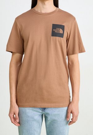 Osoba nosząca brązowy krótki T-shirt z czarnym kwadratowym logo z napisem „THE NORTH FACE” na lewej piersi, w połączeniu z jasnoniebieskimi jeansami.