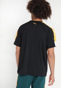 Svart idrottströja med gula detaljer på ärmarna, med rundad halsringning och en logotyp på baksidan. Lätt och andningsbart material, korta ärmar.