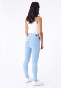 Jeans ajustados de mezclilla azul claro, diseño de cintura alta, con dos bolsillos traseros y un dobladillo limpio. Llevados con una camiseta sin mangas blanca y zapatillas.