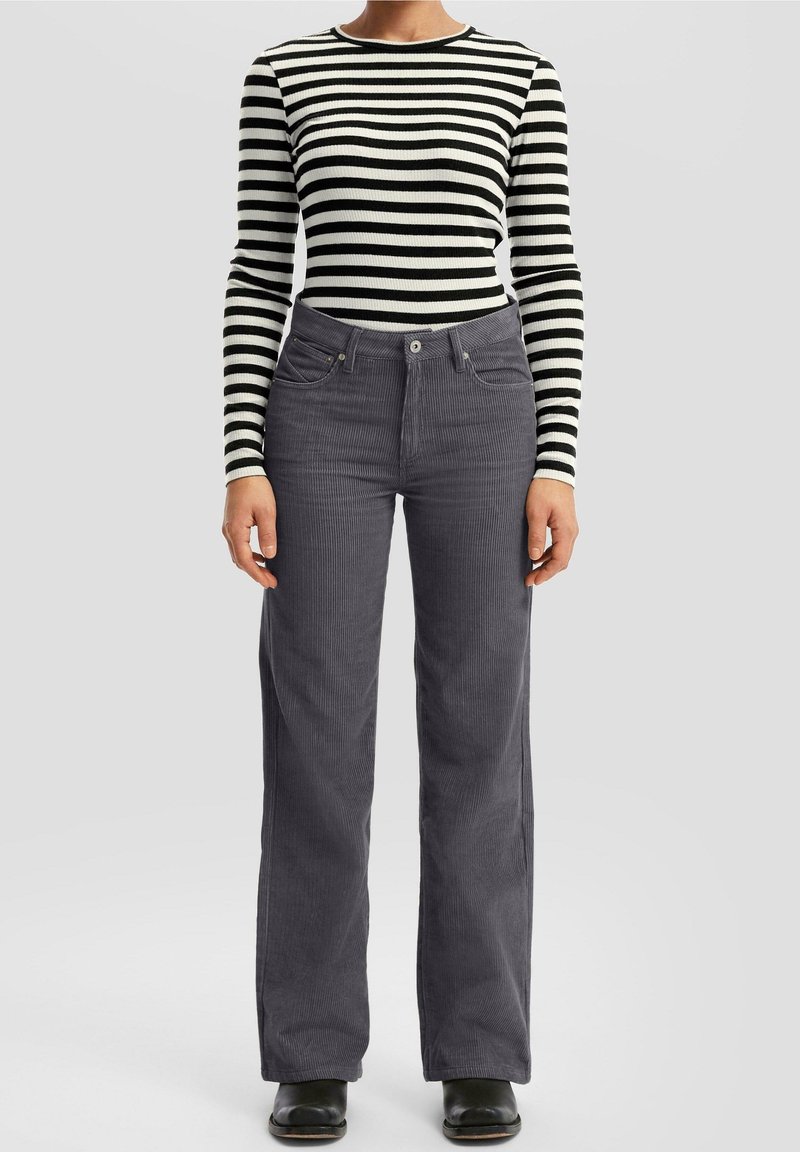 Personne portant une chemise à manches longues rayée noir et blanc, un pantalon en velours côtelé gris et des chaussures noires, debout devant un fond clair.