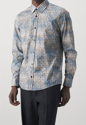 Homme portant une chemise à manches longues avec des motifs géométriques bleus et beiges et un pantalon noir sur un fond uni.