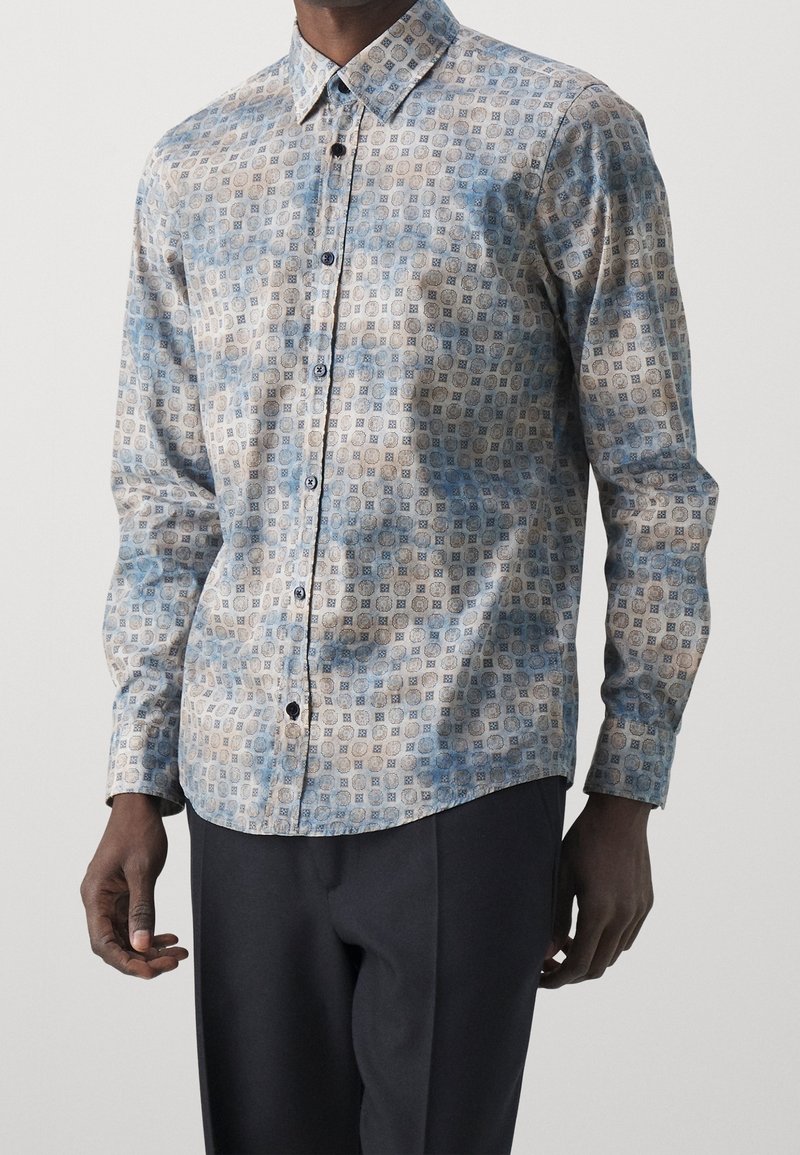 Homme portant une chemise à manches longues avec des motifs géométriques bleus et beiges et un pantalon noir sur un fond uni.
