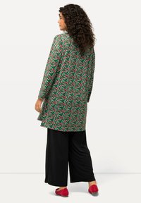 Tunica lunga con uno sfondo verde decorata con motivi circolari rossi e rosa, abbinata a pantaloni a gamba larga neri e scarpe rosse.