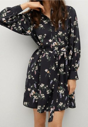 Kvinde iført en sort blomsterprintet langærmet kjole med knapper og taljebånd, poserende med den ene arm løftet ved hovedet.