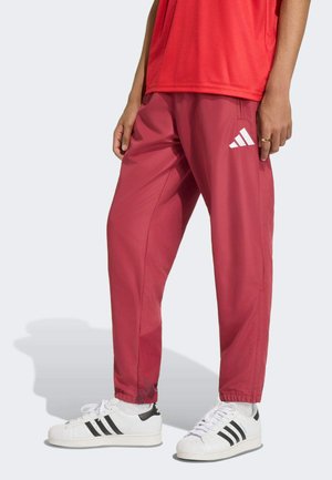 Osoba nosząca czerwone spodnie dresowe Adidas i białe buty sportowe Adidas z czarnymi paskami, stojąca na tle jednolitego jasnego tła.
