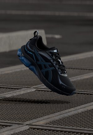 Chaussures de sport noires avec une tige en mesh respirant, des accents bleus et une semelle rembourrée. Elles disposent d'une semelle antidérapante texturée et d'une fermeture à lacets.