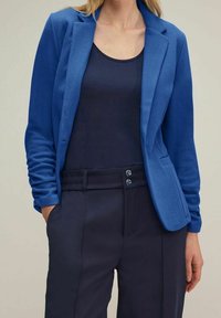 Blå kavaj i strukturerad tyg, med en knappstängning och två framfickor, bärs över en marinblå tank-top och skräddarsydda byxor.