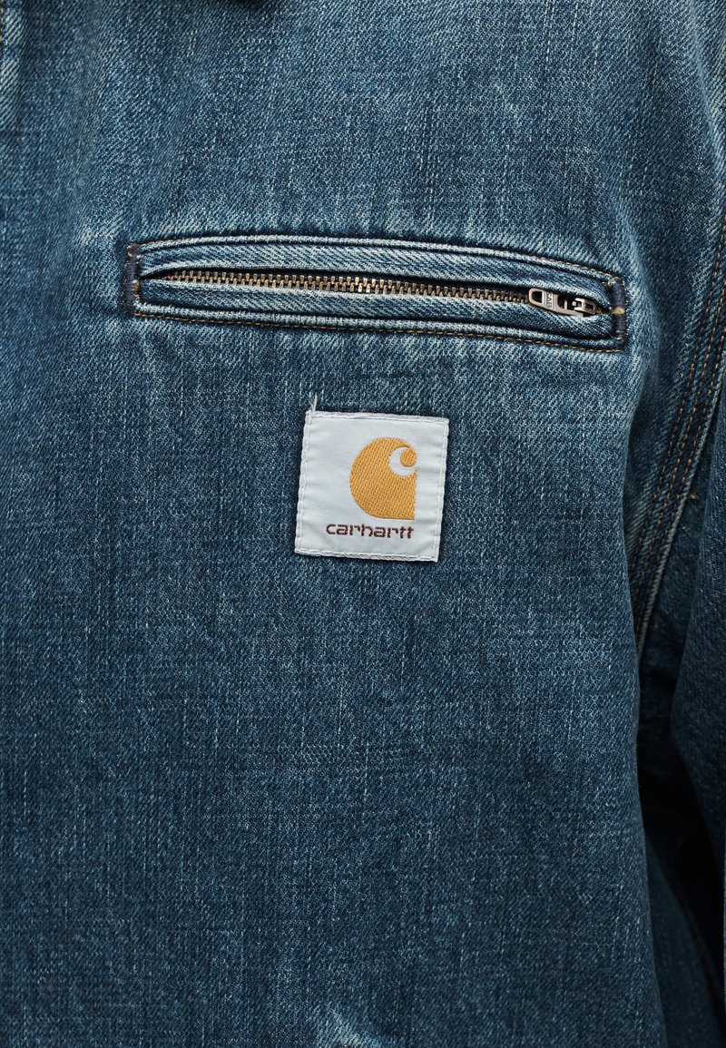 Tasca frontale della giacca di jeans con cerniera orizzontale sopra una toppa bianca con logo Carhartt con cuciture gialle e marroni.
