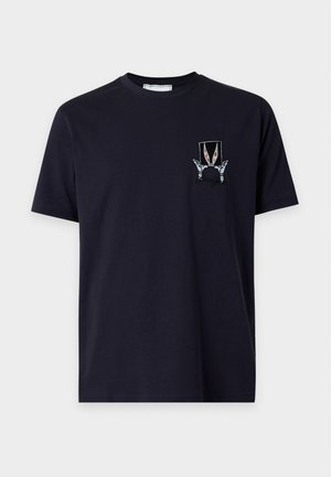 Mann trägt schwarzes Grafik-T-Shirt, lockere dunkelgraue Jeans und weiße Sneaker und steht vor weißem Hintergrund.