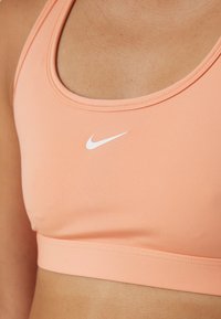 Peach sports bra with a round neckline, soft elastic band, and a white Nike logo on the front. Smooth, stretchy fabric texture.