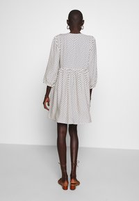 Kvinna med kort hår som bär en vit långärmad klänning med svart mönster och bruna sandaler med blockklack och remmar, stående mot en vit vägg.