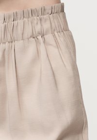 Κοντινό πλάνο σε παντελόνι με ελαστική μέση σε μπεζ απόχρωση, με λεπτή υφή, ρυτιδωτό ύφασμα και ομαλή επιφάνεια σε ανοιχτό φόντο.