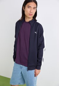 Marineblå zip-jakke med hvite sidestreker, kombinert med en lilla T-skjorte og lyse denimshorts. Har en avslappet passform og ribbestrikkede mansjetter.