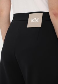 Black pants with a relaxed fit, featuring a beige label at the waist with white "MM" text. Smooth fabric, minimal design, no visible embellishments.